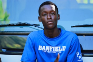 A person wearing a blue shirt with white text, sitting in front of a vehicle. The person has a neutral expression and the shirt displays the words 'BANFIELD THE PET HOSPITAL'.