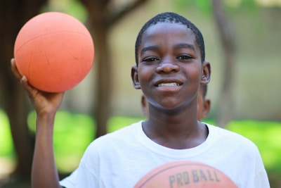 A young person is holding a basketball in one hand and smiling. The background is blurred, with hints of greenery and trees, suggesting an outdoor setting.