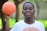 A young person is holding a basketball in one hand and smiling. The background is blurred, with hints of greenery and trees, suggesting an outdoor setting.