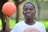 A young person is holding a basketball in one hand and smiling. The background is blurred, with hints of greenery and trees, suggesting an outdoor setting.