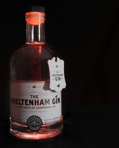 A bottle of gin with a detailed label featuring the words 'The Cheltenham Gin' and 'The Taste of Champions'. The bottle cap and neck are illuminated with a red glow, and a tag is attached around the neck.
