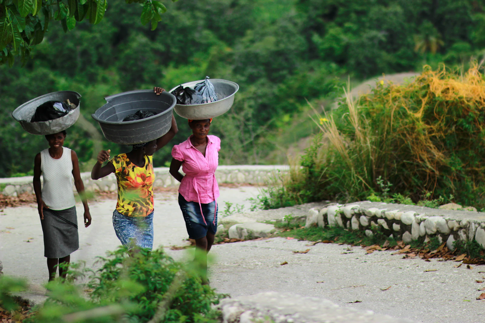 Three women walk together carrying large water containers balanced on their heads along a dirt path.