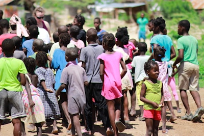 A group of children and a few adults gathered outdoors in a lively and colorful setting. The children are wearing a variety of bright, casual clothing and appear to be engaged in an outdoor activity on a sunny day. In the background, some greenery and a few structures are visible, suggesting a rural or suburban environment.