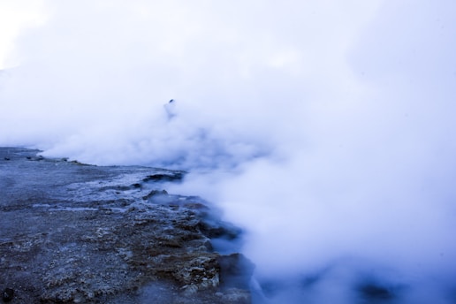 Thick clouds of steam or mist rise from a rocky surface, creating an ethereal and mysterious atmosphere. The ground appears rugged and uneven, with the steam obscuring most of the background details.