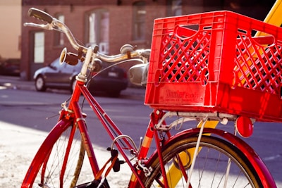A red bicycle with curved handlebars is parked on a street. Attached to the back of the bike is a red plastic crate, likely used for carrying items. The sunlight casts shadows on the bike and crate, highlighting their bright colors. In the background, there is a parked car and a brick building.