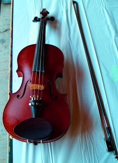 A violin with a rich red-brown color is placed on a white cloth. The violin's body, strings, and fine tuners are clearly visible. Next to the violin, there is a bow lying parallel to it.
