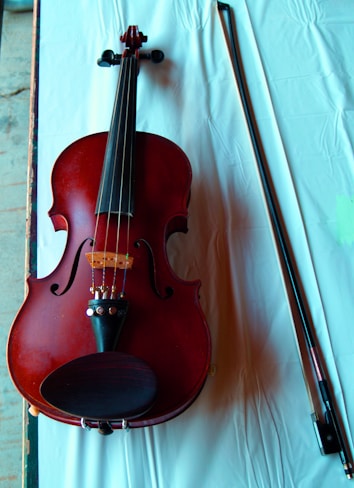 A violin with a rich red-brown color is placed on a white cloth. The violin's body, strings, and fine tuners are clearly visible. Next to the violin, there is a bow lying parallel to it.
