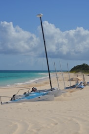 Sailboats are lined up on a sandy beach, with turquoise ocean waters gently lapping at the shore. The sky is bright with white, fluffy clouds scattered across it. In the background, lush greenery contrasts with the beige sands.