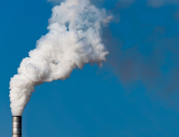 A chimney releasing a large plume of white smoke against a clear blue sky. The smoke appears thick and voluminous, suggesting industrial activity.