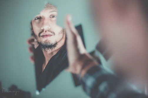 A person surrounded by judging faces with a broken mirror nearby, conveying social pressure and strong psychological emotion in a modern dark style.