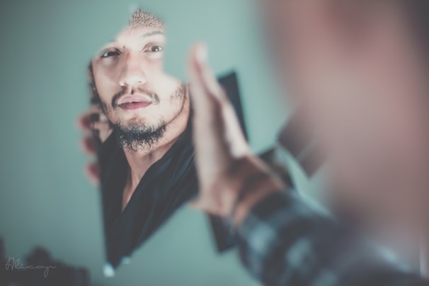 A man's face is reflected in a broken piece of mirror held in their hand. The focus is on their thoughtful expression, with soft lighting and a blurred background enhancing the introspective mood.