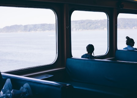 Inside a transport vehicle with large windows overlooking a body of water, two people are seated, facing the view. The interior appears spacious with blue seating, and the atmosphere is calm and quiet.
