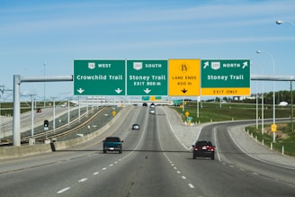 A multi-lane highway with several overhead signs indicating directions for Crowchild Trail and Stoney Trail in both north and south directions. The road has vehicles traveling on it with clear skies and some grassy areas on the side.