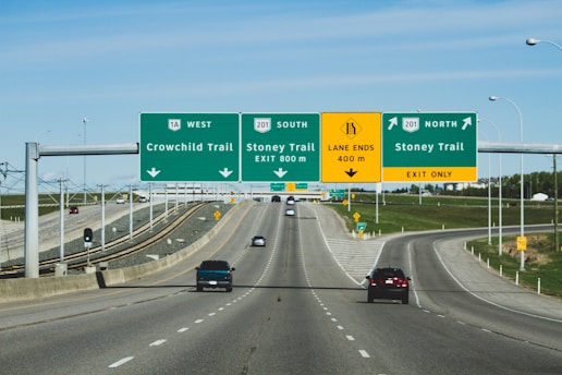 A multi-lane highway with several overhead signs indicating directions for Crowchild Trail and Stoney Trail in both north and south directions. The road has vehicles traveling on it with clear skies and some grassy areas on the side.