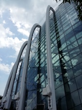 A modern glass building features large, curved arches that create a distinctive architectural design. The building's reflective glass panels mirror the cloudy sky and surrounding environment, adding depth and dynamism to the structure.