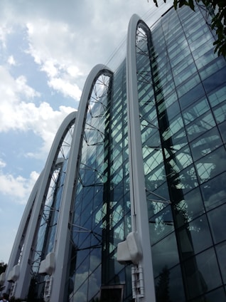 A modern glass building features large, curved arches that create a distinctive architectural design. The building's reflective glass panels mirror the cloudy sky and surrounding environment, adding depth and dynamism to the structure.