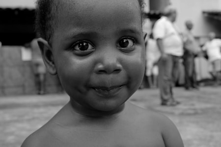 A close-up of a young child's face with wide, curious eyes and a slight smile. The background is blurred, showing several adults standing and interacting in an outdoor setting. The image is in black and white, emphasizing the child's expression.