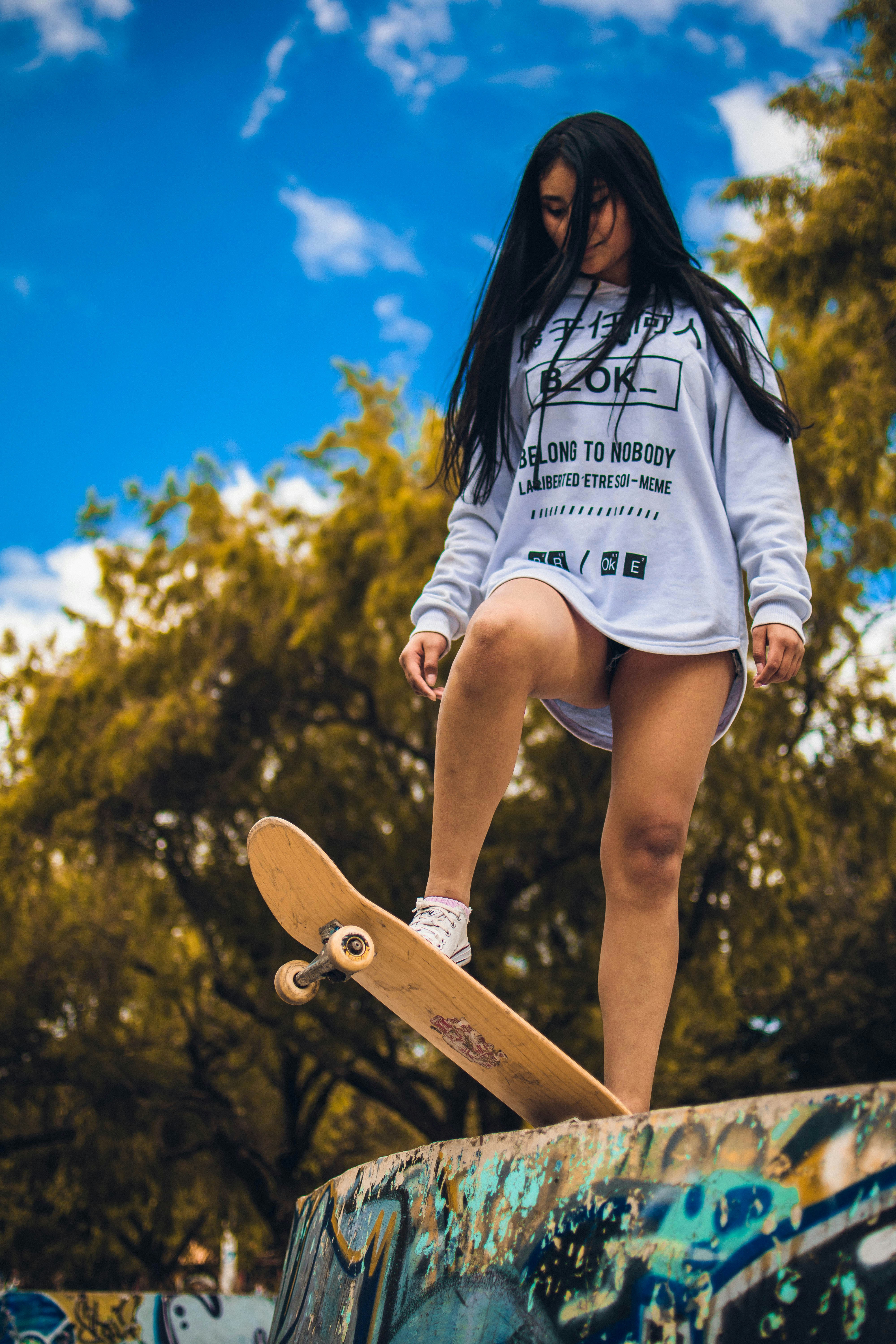 A person stands confidently on a skateboard positioned at the edge of a graffiti-covered ramp, with long hair blowing slightly in the wind. They are wearing a white sweatshirt with black text and graphics, paired with white sneakers. The background features lush green trees under a bright blue sky with scattered clouds.