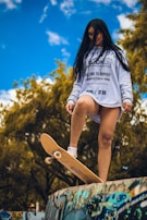 A person stands confidently on a skateboard positioned at the edge of a graffiti-covered ramp, with long hair blowing slightly in the wind. They are wearing a white sweatshirt with black text and graphics, paired with white sneakers. The background features lush green trees under a bright blue sky with scattered clouds.