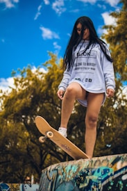 A person stands confidently on a skateboard positioned at the edge of a graffiti-covered ramp, with long hair blowing slightly in the wind. They are wearing a white sweatshirt with black text and graphics, paired with white sneakers. The background features lush green trees under a bright blue sky with scattered clouds.