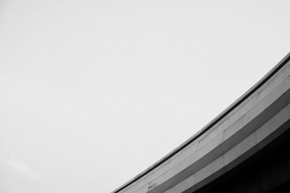A minimalist black and white photograph featuring a curved architectural element against a vast, empty sky. The structure has a sleek and modern design, with clean lines and smooth surfaces.