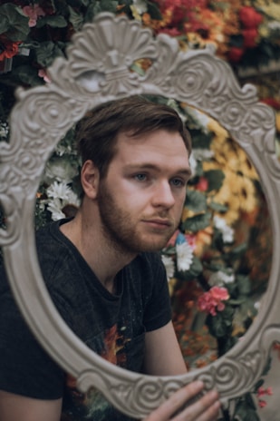 A person with short hair gazes through an ornate, oval mirror. In the background, a variety of colorful, lush flowers including red, yellow, and green fill the scene, creating a vibrant and artistic atmosphere. The person appears thoughtful, adding a contemplative quality to the image.