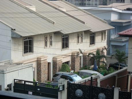 Row of modern townhouses with beige walls and slanted metal roofs, surrounded by a gated fence. The front yards have various plants and trees, and multiple satellite dishes are attached to the building. Cars are parked in the driveway.
