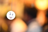 A smiley face emoticon is seen hanging from a string, with a blurred background that has warm, golden tones. The emoticon has large, expressive eyes and an open mouth with a tongue sticking out.