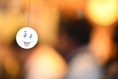 A smiley face emoticon is seen hanging from a string, with a blurred background that has warm, golden tones. The emoticon has large, expressive eyes and an open mouth with a tongue sticking out.