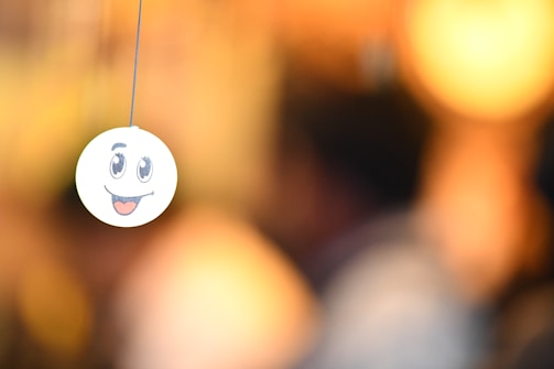 A smiley face emoticon is seen hanging from a string, with a blurred background that has warm, golden tones. The emoticon has large, expressive eyes and an open mouth with a tongue sticking out.