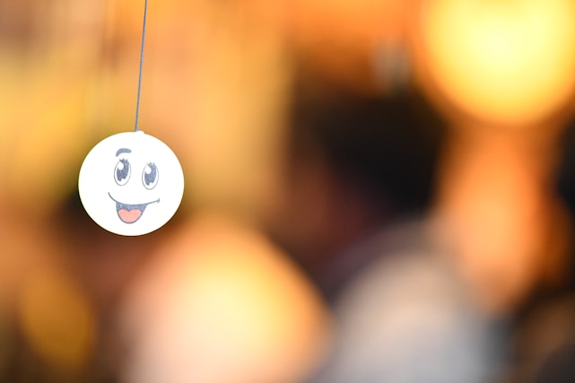 A smiley face emoticon is seen hanging from a string, with a blurred background that has warm, golden tones. The emoticon has large, expressive eyes and an open mouth with a tongue sticking out.