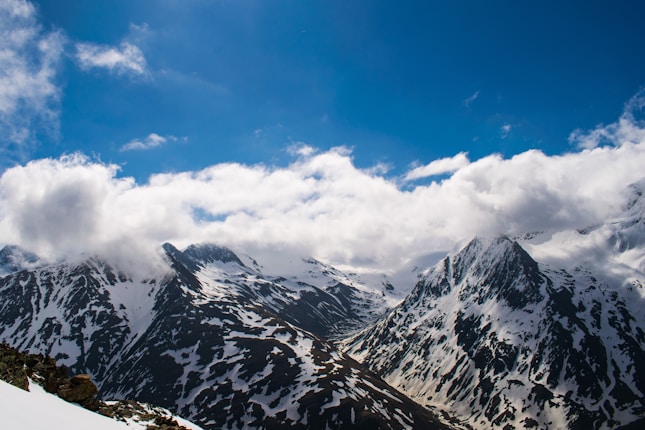 Snow-covered mountains under a clear blue sky, with patches of clouds floating above the peaks.