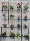 A decorative arrangement of multiple flower pots is mounted on an iron grid against a textured white wall. Each pot contains vibrant artificial flowers in various colors and patterns, including stripes and polka dots. The pots are arranged in a neat, organized fashion, creating a visually pleasing geometric pattern.