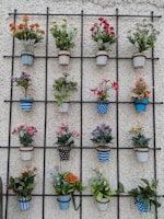 A decorative arrangement of multiple flower pots is mounted on an iron grid against a textured white wall. Each pot contains vibrant artificial flowers in various colors and patterns, including stripes and polka dots. The pots are arranged in a neat, organized fashion, creating a visually pleasing geometric pattern.