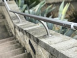 A concrete staircase is bordered by a stone wall with a sleek metallic handrail. In the background, there are large, pointed green leaves from plants, possibly succulents, providing contrast to the stone and metal structure.