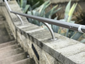 A concrete staircase is bordered by a stone wall with a sleek metallic handrail. In the background, there are large, pointed green leaves from plants, possibly succulents, providing contrast to the stone and metal structure.