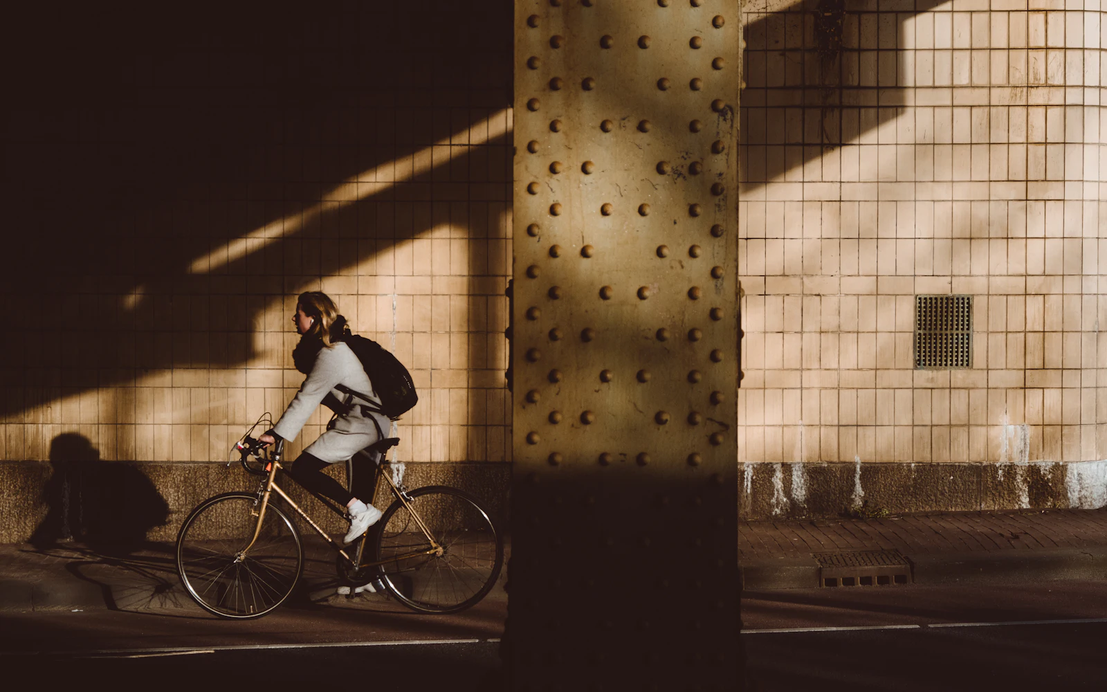 A rider with a backpack cycling past a riveted steel pillar, moving from sunlight into shadow