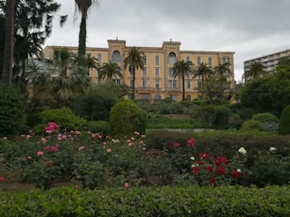 An elegant multi-story building with a classical design is surrounded by lush greenery and a variety of colorful flowers. Tall palm trees and other vegetation create a serene garden atmosphere.