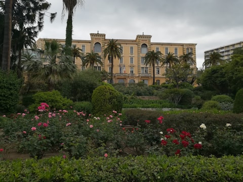 An elegant multi-story building with a classical design is surrounded by lush greenery and a variety of colorful flowers. Tall palm trees and other vegetation create a serene garden atmosphere.