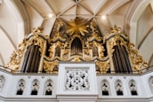 An ornate pipe organ with intricate golden details and decorative elements is set against a background of a vaulted ceiling. The structure features multiple tall pipes, embellished carvings, and a large golden starburst design in the center.