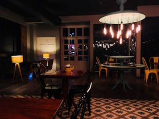 A dimly lit, cozy indoor space with wooden tables and chairs. Several pendant lights hang from the ceiling, casting a warm glow. On the left, lamps with fabric shades are placed near a wall with shutters, adding to the ambient lighting. Yellow metal chairs provide a pop of color against the darker furniture, and patterned tiles cover the floor.