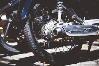 Close-up view of a motorcycle wheel and exhaust system. The focus is on the tire tread, spokes, and metallic components, such as the brake disc and shiny exhaust pipe. Sunlight casts contrasting shadows on the concrete ground.