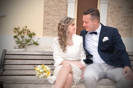 A couple is sitting on a wooden bench, smiling at each other. The woman is wearing a white dress and holding a bouquet of flowers with yellow and white blooms. The man is dressed in a formal blue jacket and beige trousers. The background consists of a textured wall and a potted plant.