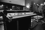 A vintage electric piano sits prominently in a music studio. The setting includes microphones, a lamp, and various recording equipment scattered throughout the room. The space has a classic, slightly cluttered appearance, indicative of a working studio environment.