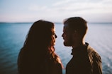 A couple stands close together, silhouetted against a backdrop of calm water and a partly cloudy sky. The sunlight creates a warm glow around their faces as they gaze into each other's eyes.