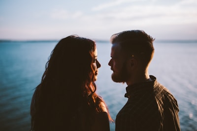 A couple stands close together, silhouetted against a backdrop of calm water and a partly cloudy sky. The sunlight creates a warm glow around their faces as they gaze into each other's eyes.