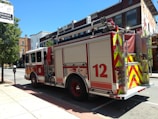 A fire truck is parked on a street in an urban setting. The vehicle has a red and white color scheme with the number 12 prominently displayed on its side. Nearby, there are buildings with brick facades and a tree providing some greenery. A catering sign is visible above the sidewalk.