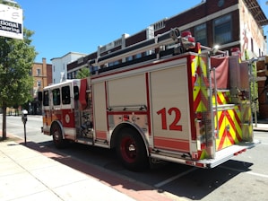 A fire truck is parked on a street in an urban setting. The vehicle has a red and white color scheme with the number 12 prominently displayed on its side. Nearby, there are buildings with brick facades and a tree providing some greenery. A catering sign is visible above the sidewalk.