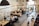 A spacious, modern office setting with several people working at large wooden desks. Overhead string lights add a warm ambiance, and there are computers and office supplies on the desks. Large windows allow natural light to enter, and there are potted plants in the background.