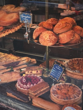 A variety of baked goods are displayed in a bakery window, including loaves of bread, pastries, and desserts. There are neatly arranged gougères, layered cakes topped with raspberries, and fruit tarts.